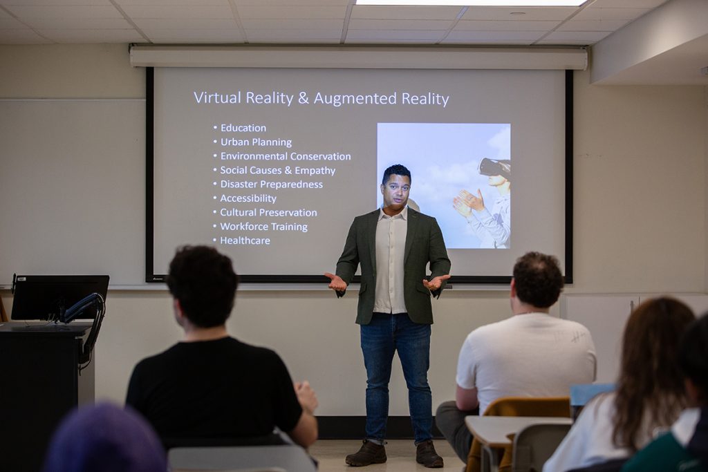 John Christensen, associate professor of communication, gestures and speaks in front of a PowerPoint presentation while teaching a class.