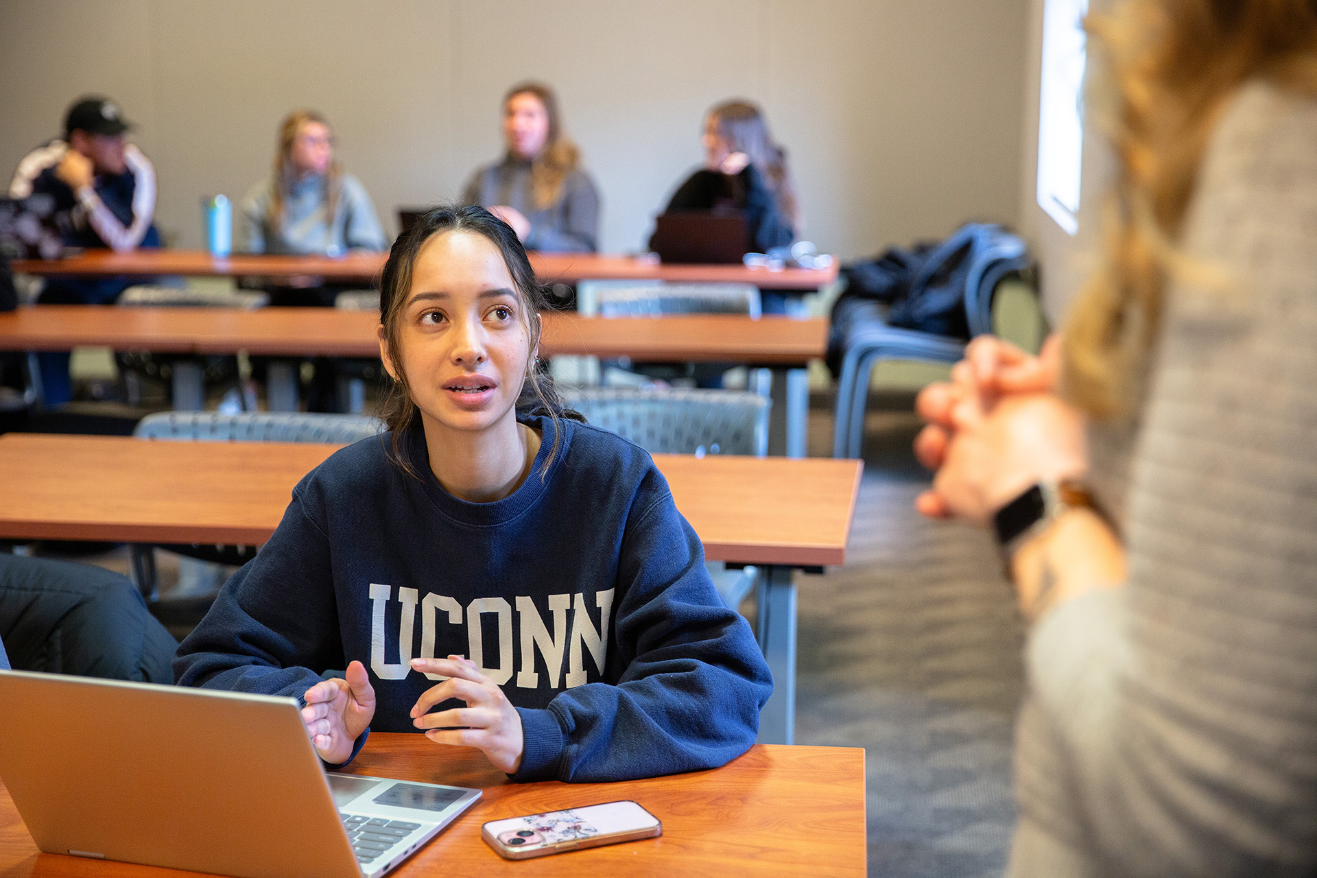 A student wearing a UConn sweatshirt asks their professor a question while gesturing to a laptop.